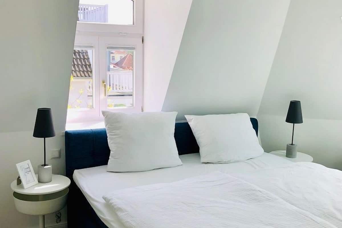 A bedroom with a bed, two bedside tables, a lamp on each table, a window, and a view of buildings outside.