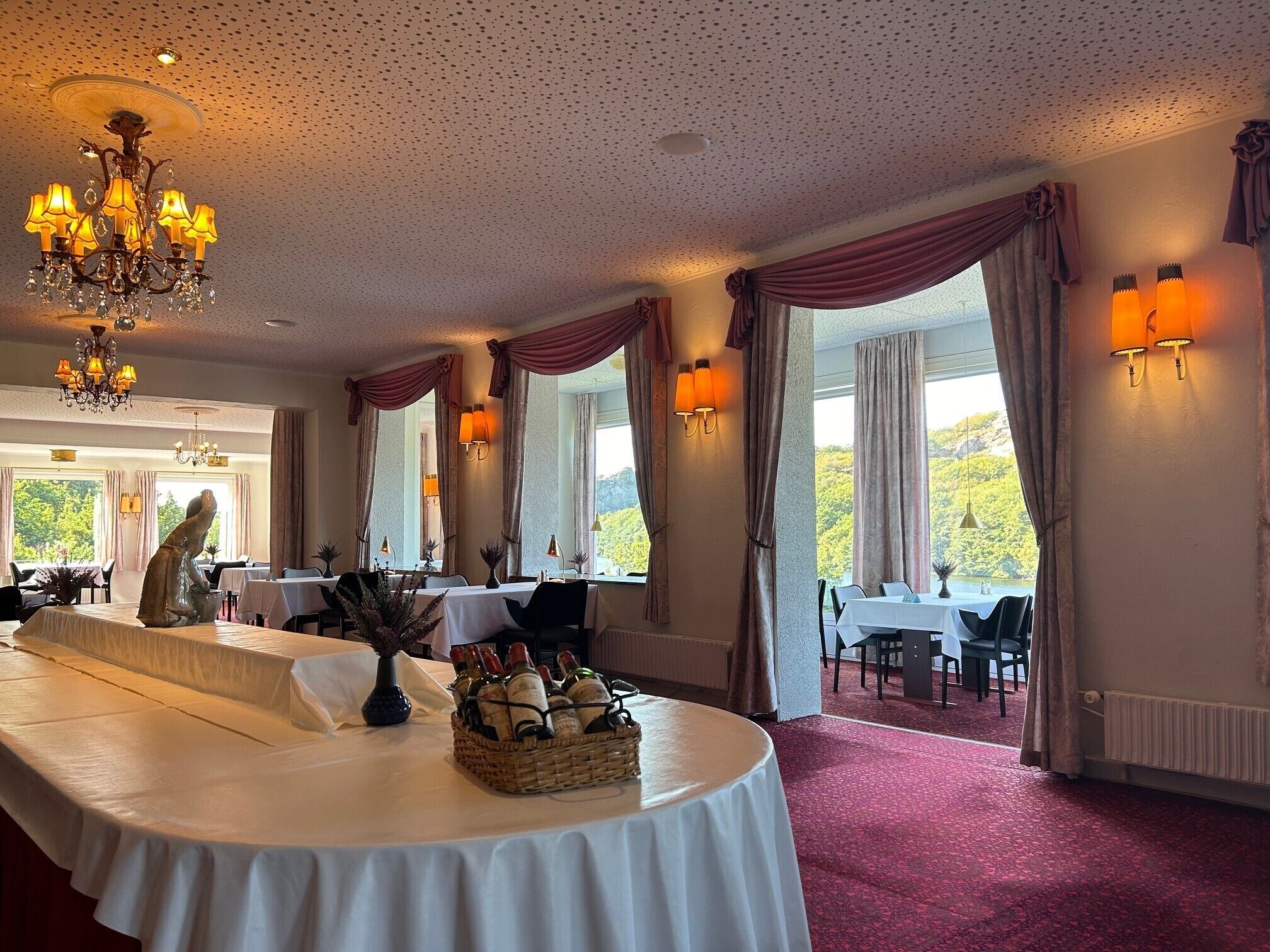 Une salle à manger avec des tables dressées pour le service, un lustre et de grandes fenêtres ornées de rideaux.