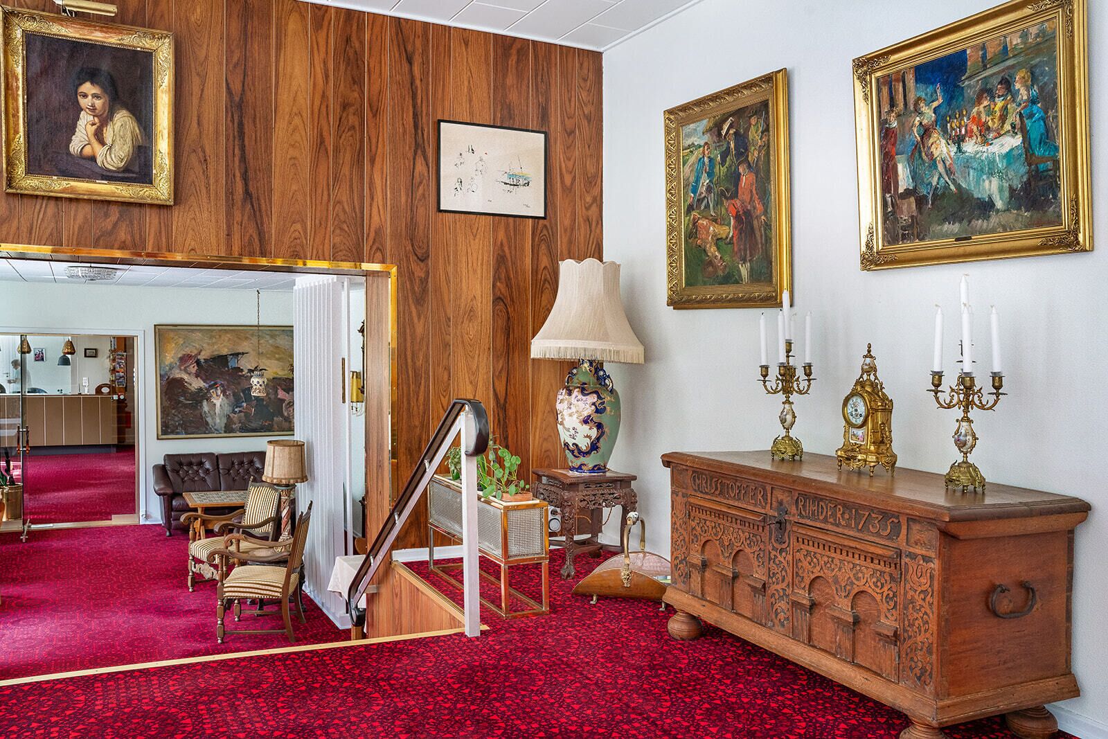 A room with a red carpet, wooden panel walls, a large wooden cabinet with candelabras, a vintage lamp, and framed artwork.