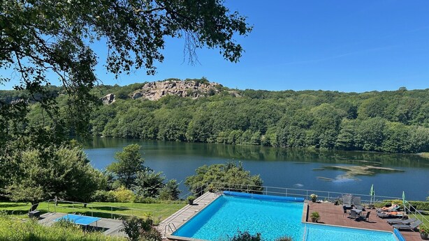 A rectangular swimming pool with a clear blue water, surrounded by a deck with lounge chairs and umbrellas, overlooking a lake with a forested hill in the background. - Hotel Hammersø Apartments - Adults Only (Allinge)