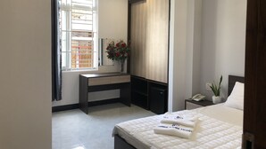 A modern hotel room with a bed, bedside table, and a desk with a chair. There is a window with curtains, a plant, and a mirror on the wall. - Hotel Hong Phuc (Quy Nhon Nam)