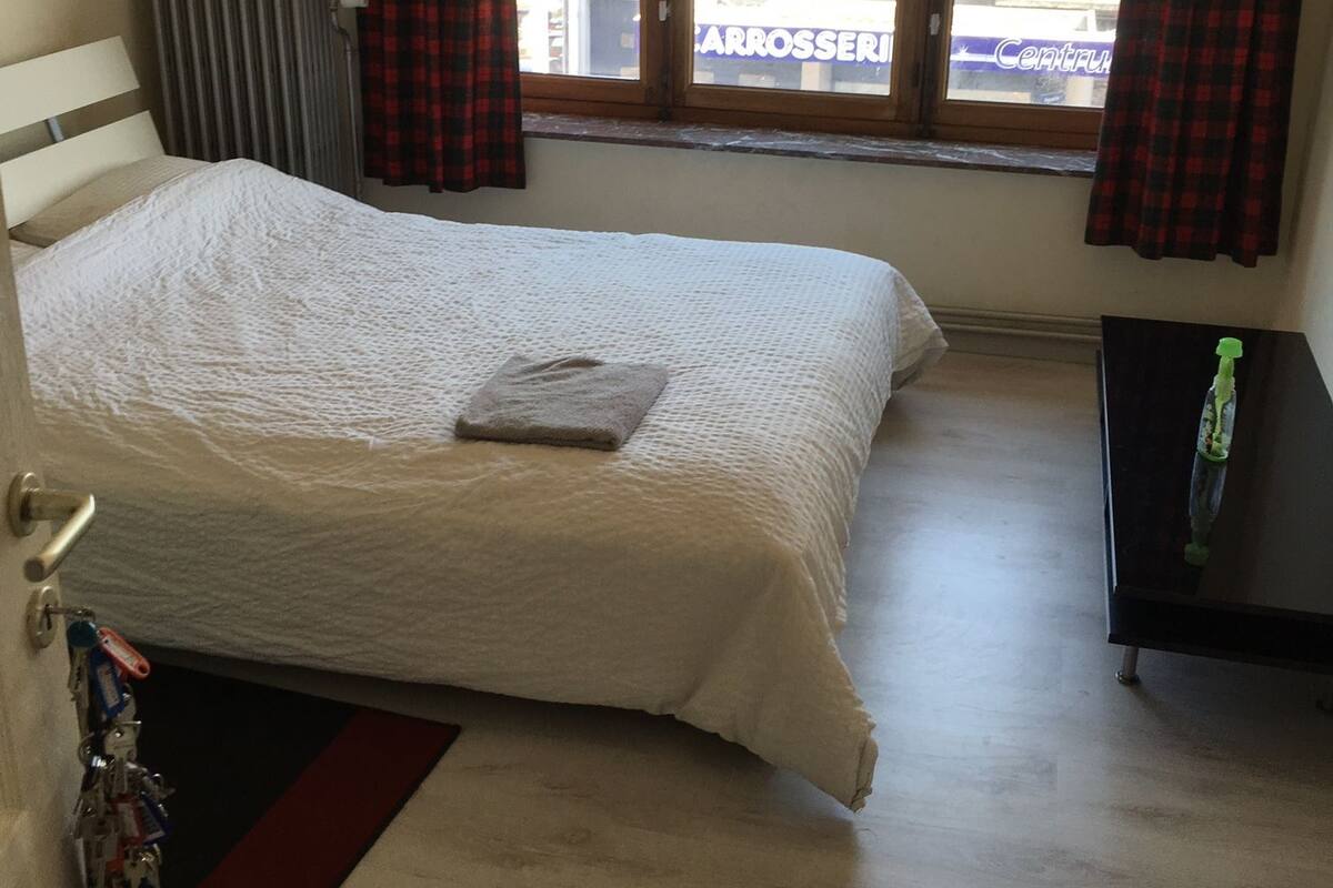 Een slaapkamer met een bed, een radiator, een raam met gordijnen, een klein tafeltje en een deur.