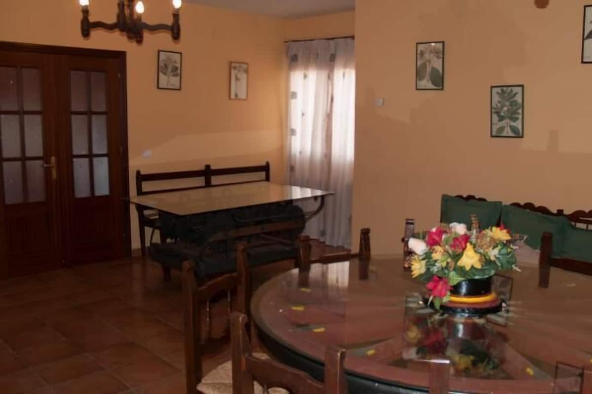 Un comedor con una mesa redonda, sillas, un jarrón de flores y cuadros en la pared.
