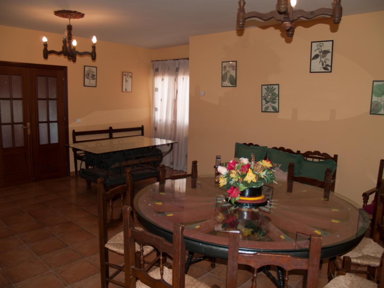 A dining area with a round glass table surrounded by wooden chairs. There is a chandelier above the table and a bench along the wall.