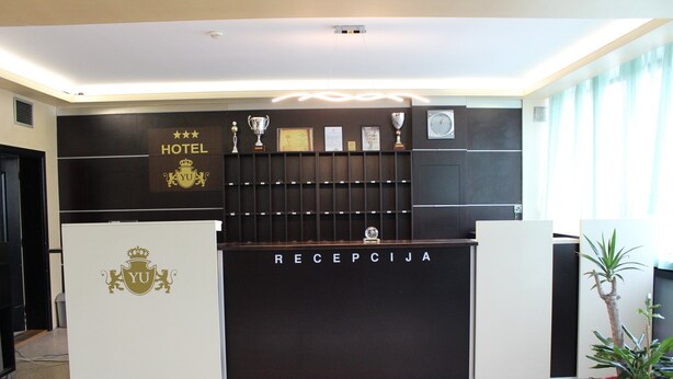 A hotel reception desk with a dark wood finish, a white reception area, and a reception sign. - Hotel Yu (East Sarajevo)