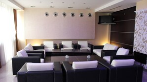 A modern lounge area with black and white sofas arranged around a central coffee table, a large flat-screen TV mounted on the wall, and a patterned curtain on the right. - Hotel Yu (East Sarajevo)