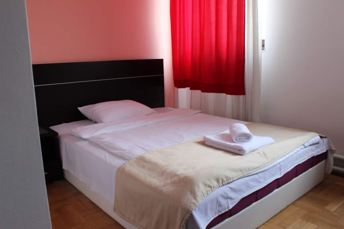 A neatly made bed with white linens, a beige blanket, and a rolled towel.