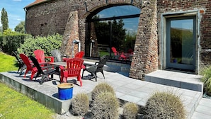 A brick building with a tiled roof, outdoor seating with red and black chairs, and a glass door leading to a patio area. - Gîte Les Oliviers – 4 people – 2 bedrooms – 1 bathroom – Indoor pool (Arques)