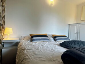 Ein ordentlich bezogenes Bett mit weißer Daunendecke und zwei gemusterten Kissen, eine Nachttischlampe und ein Kleiderschrank in einem schlichten Zimmer.