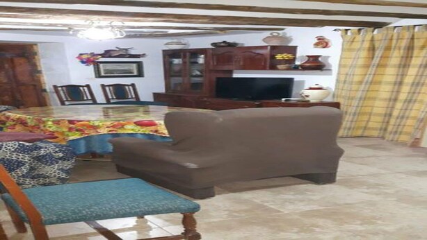 Un coin repas avec une table recouverte d’une nappe colorée, un fauteuil marron et une chaise bleue. On aperçoit un téléviseur posé sur un meuble en bois, ainsi que divers objets décoratifs accrochés au mur.