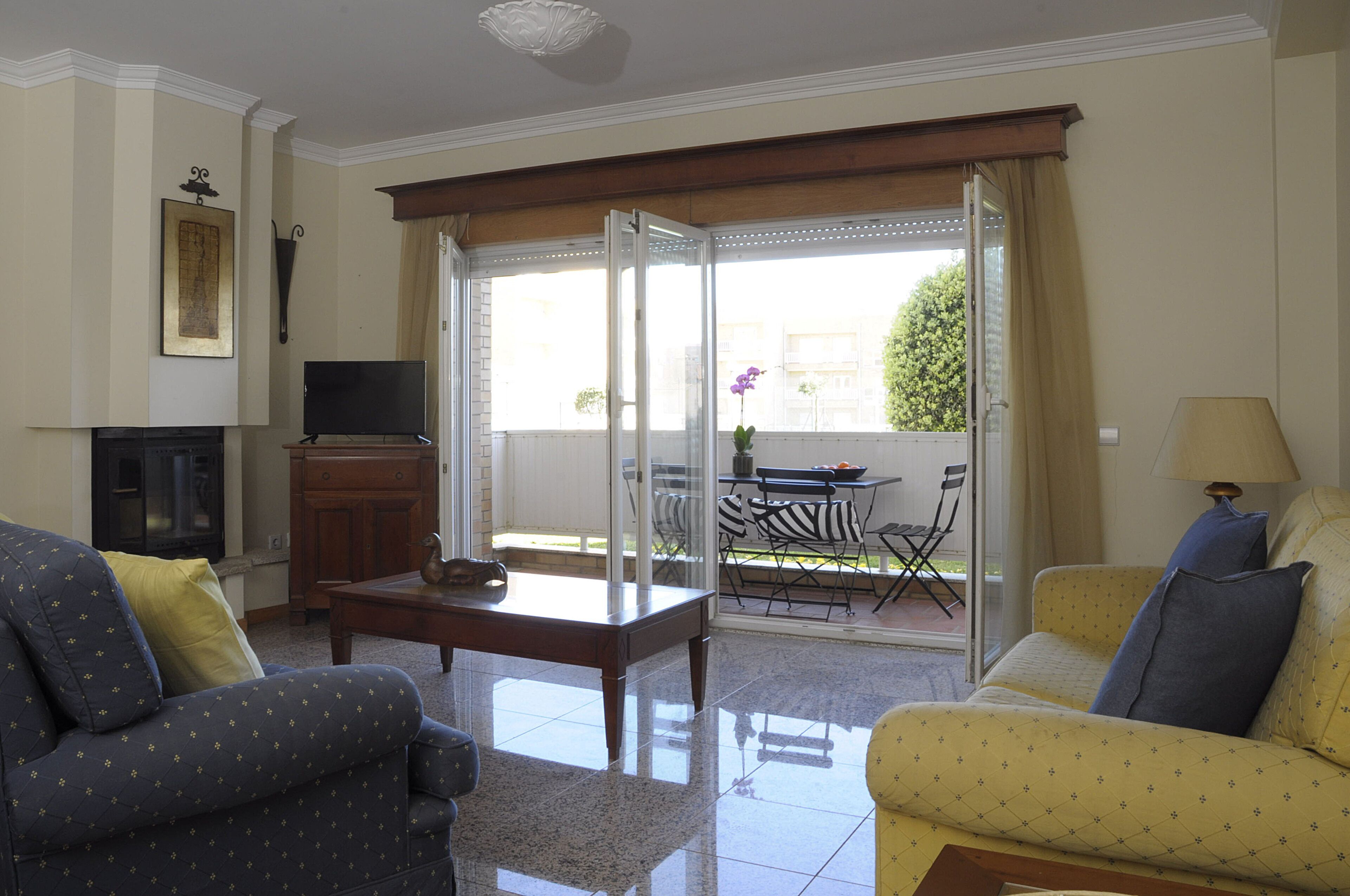 A living room with a sofa, armchair, TV, fireplace, and a view of an outdoor patio with a table and chairs.