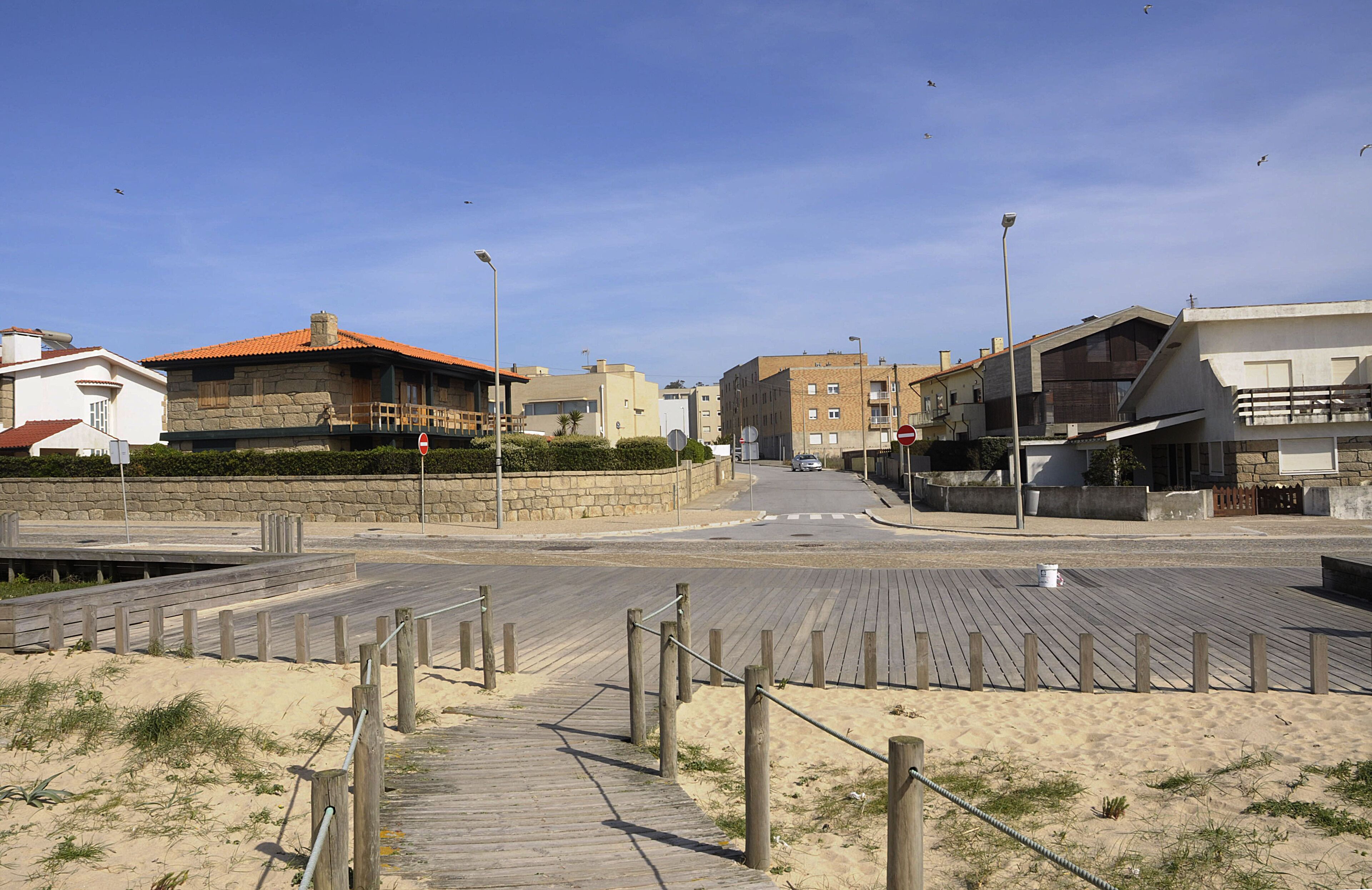 A coastal street with a wooden boardwalk leading to a sandy beach, surrounded by residential buildings.
