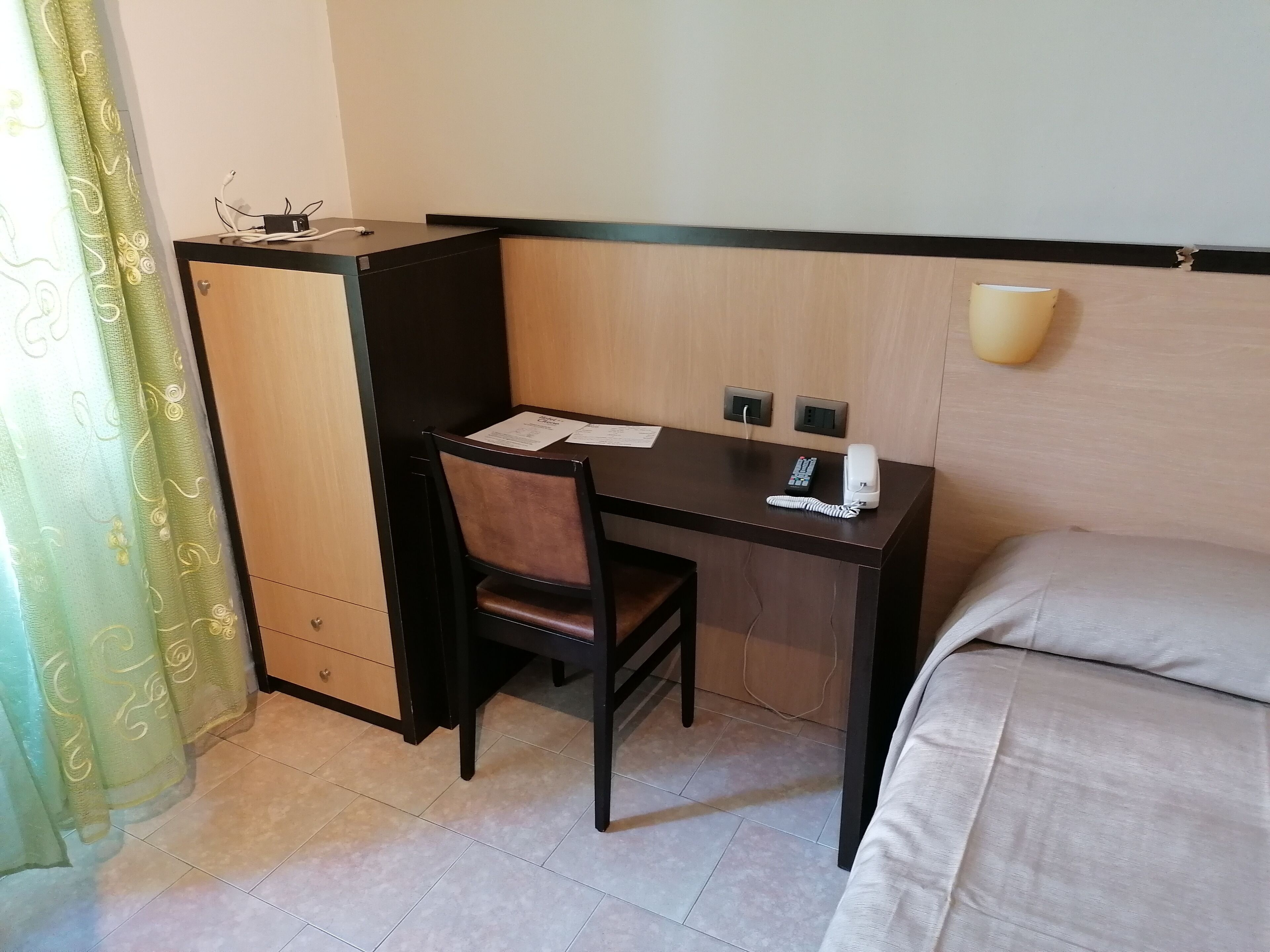 Ein kleines Hotelzimmer mit Schreibtisch, Stuhl und Bett. Auf dem Schreibtisch befinden sich ein Telefon und eine Steckdose.