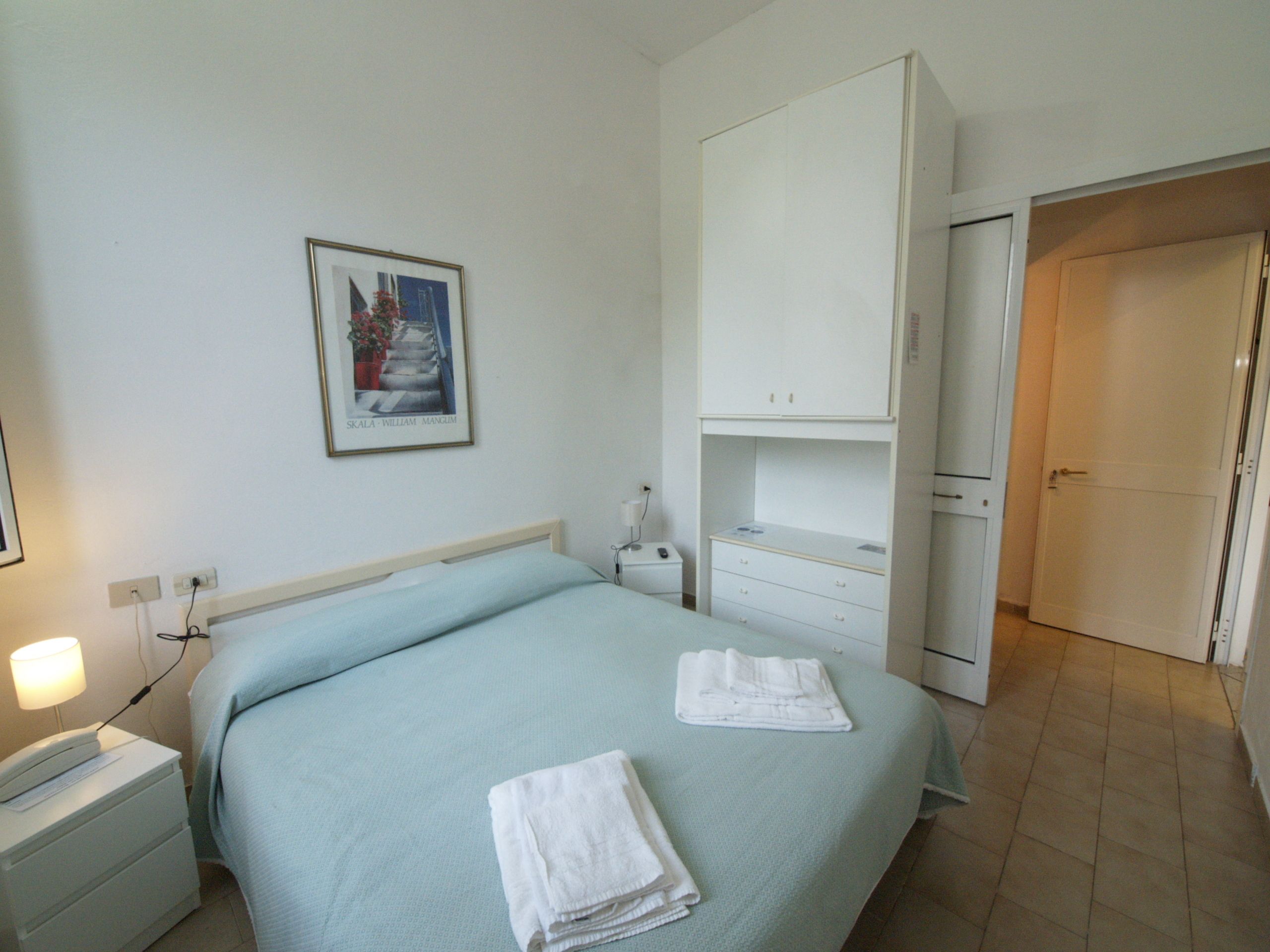 A hotel room with a bed, a nightstand, a desk, a wardrobe, and a framed picture on the wall.