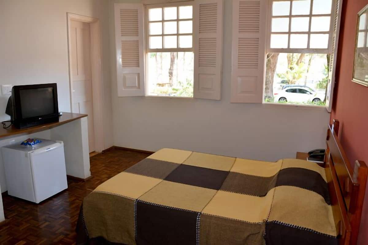 A room with a bed, a small desk with a television, a refrigerator, and windows with shutters.