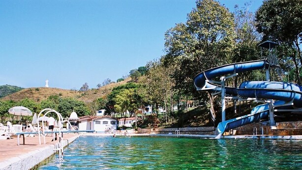A swimming pool with clear water, a water slide, and a hill with a statue in the background. - Hotel Termópolis (Sao Sebastiao do Paraiso)
