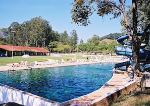 A swimming pool with lounge chairs and a water slide, surrounded by trees and a building with a red roof. - Hotel Termópolis (Sao Sebastiao do Paraiso)