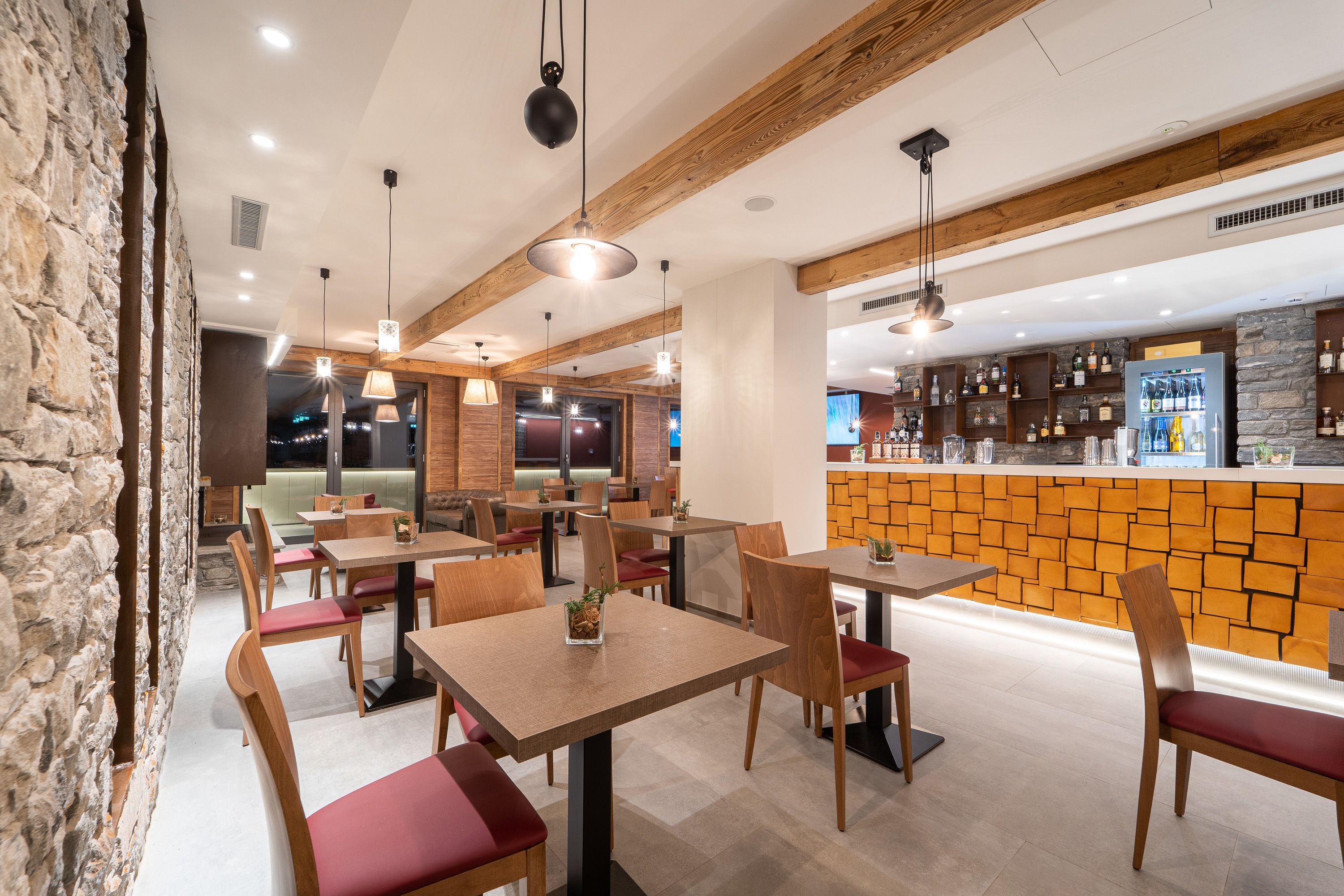 Un restaurante con mesas y sillas de madera, una pared de piedra y una zona de bar con estantes llenos de botellas.