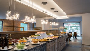A buffet area with a variety of food options, a stone counter, hanging pendant lights, and a view of the outdoors through large windows. - TH Courmayeur (Courmayeur)