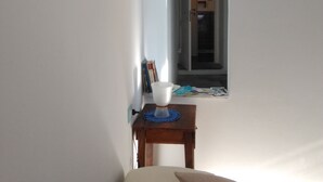 Un pasillo estrecho con una cama al final, una mesita con una lámpara y una puerta que da a otra habitación.