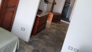 Una habitación con un armario de madera, un pequeño mueble de madera, suelo de baldosas y un acceso a un baño con ducha y aire acondicionado.