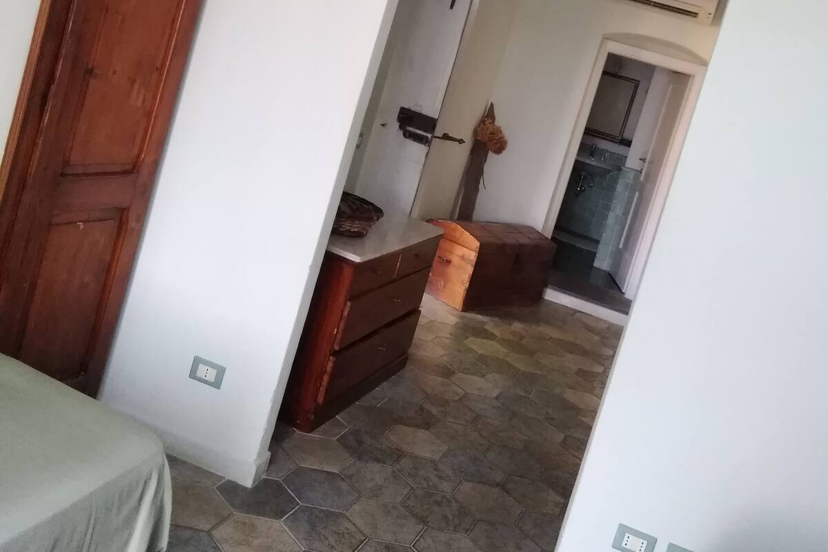 A room with a wooden wardrobe, a small wooden cabinet, a tiled floor, and a doorway leading to a bathroom with a shower and an air conditioning unit.