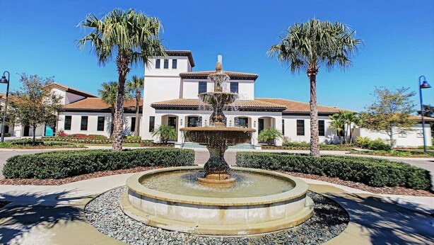A fountain with a tiered design, surrounded by a circular stone border. In the background, there is a two-story building with a tiled roof and a white facade. Palm trees are visible around the fountain.