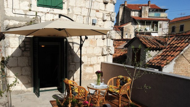 Un pequeño espacio al aire libre con dos sillas de mimbre, una mesa y un paraguas color beige. Hay plantas en macetas y una mesa pequeña con un jarrón de flores. El área está junto a un edificio de piedra con tejado de baldosas.