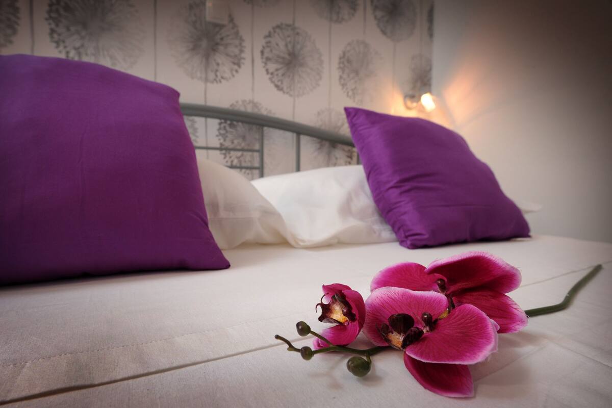 Un lit avec des oreillers violets et blancs, une orchidée rose et un mur orné d’un motif floral.