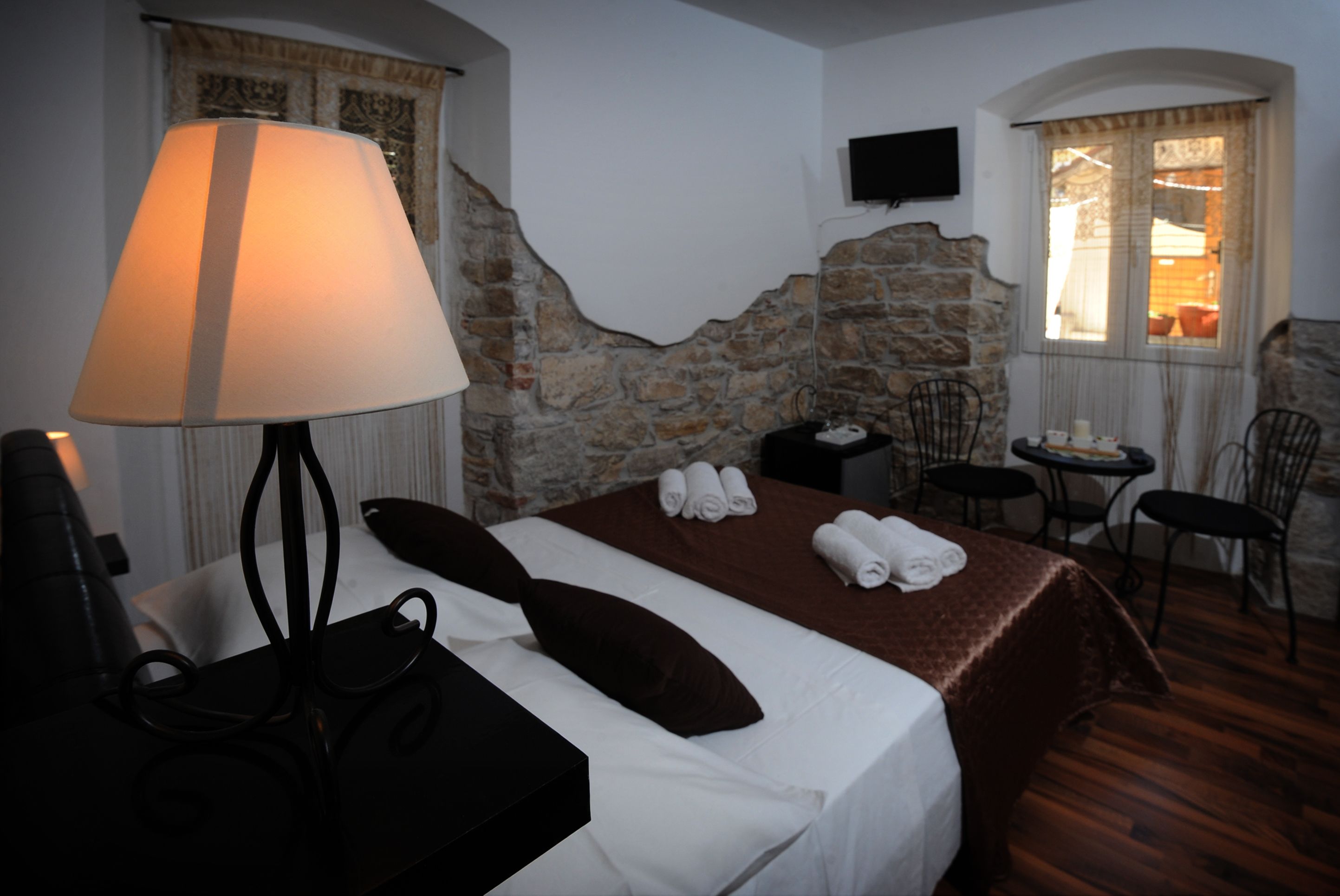 A hotel room with a bed, a desk with a lamp, a chair, a small table, and a stone wall.