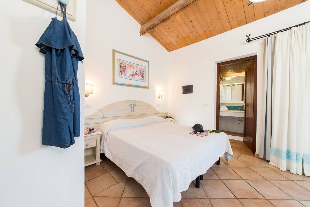 Een slaapkamer met een bed, een nachtkastje, een blauw kledingstuk dat hangt, een ingelijste foto, een badkamer met een spiegel en een deur.