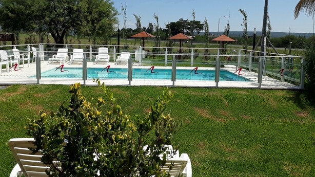 Une piscine entourée d’une clôture et de verdure, avec des chaises longues blanches et des parasols.