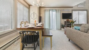 A dining area with a wooden table, chairs, and a chandelier. There is a television mounted on the wall and a couch in the living area.
