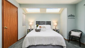 A bedroom with a large bed, two bedside tables, a chair, a skylight, and a wooden door.