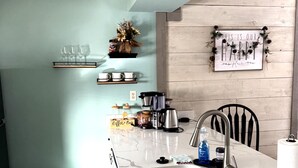 A kitchen with a white marble countertop, a stainless steel sink, and a wooden backsplash. There are shelves with kitchenware and a coffee bar sign.