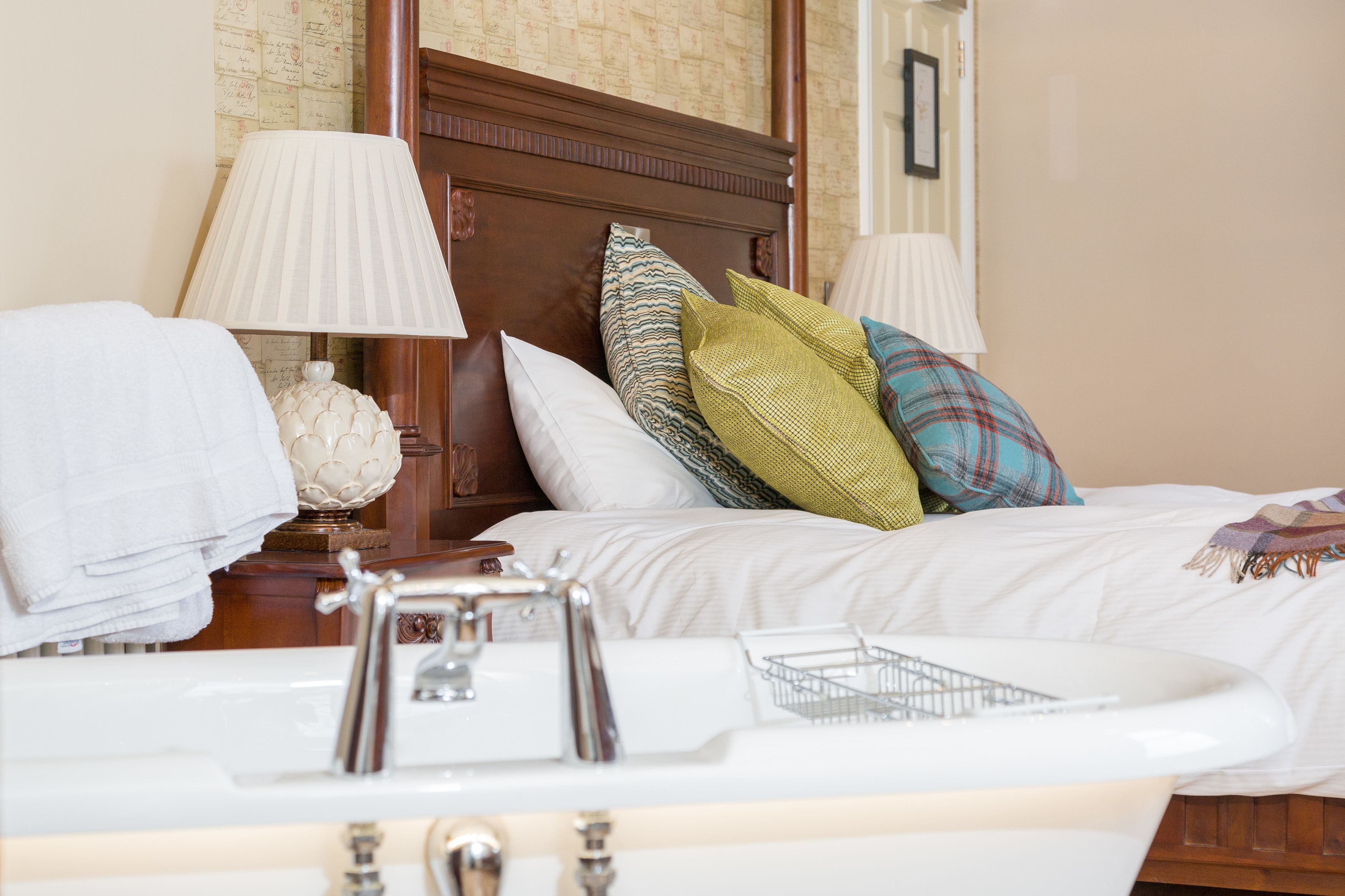 Una habitación de hotel con cama, dos lámparas de noche, una mesita de noche con lámpara, un objeto decorativo y un baño con lavabo y espejo.
