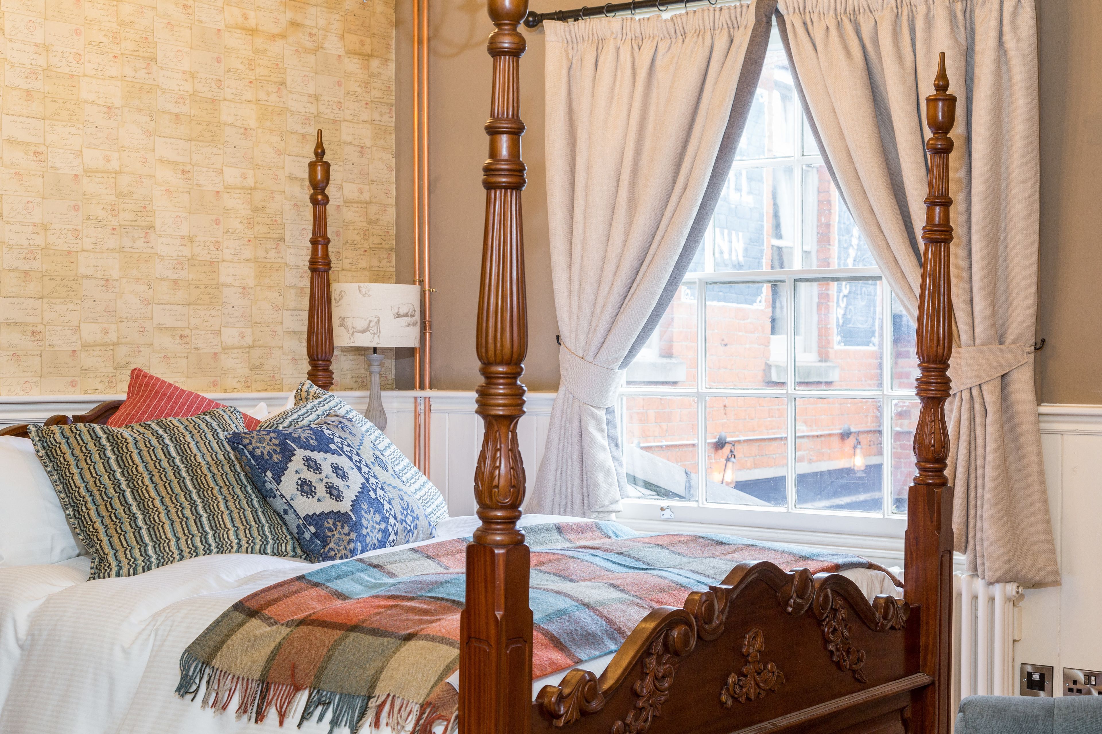 Una cama con dosel, con colcha escocesa, varios cojines estampados y una ventana con cortinas.