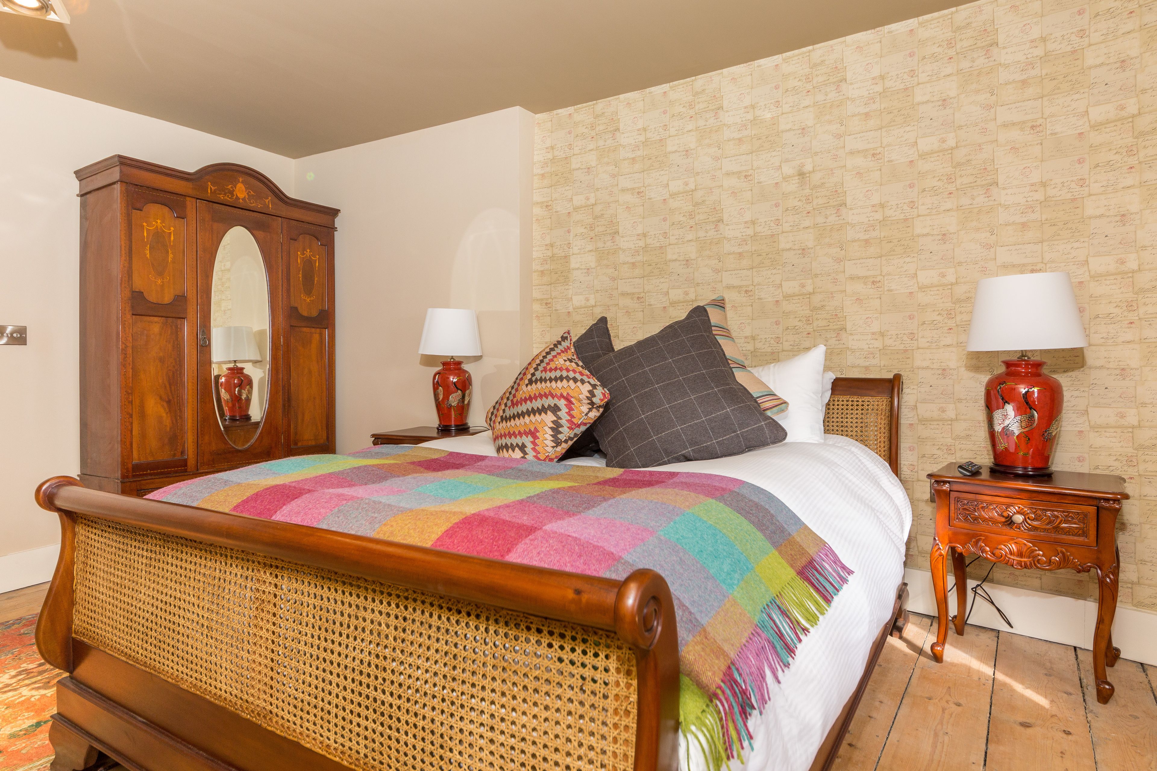 Un dormitorio con cama de madera, un espejo grande, dos mesitas de noche con lámparas y un papel tapiz estampado.