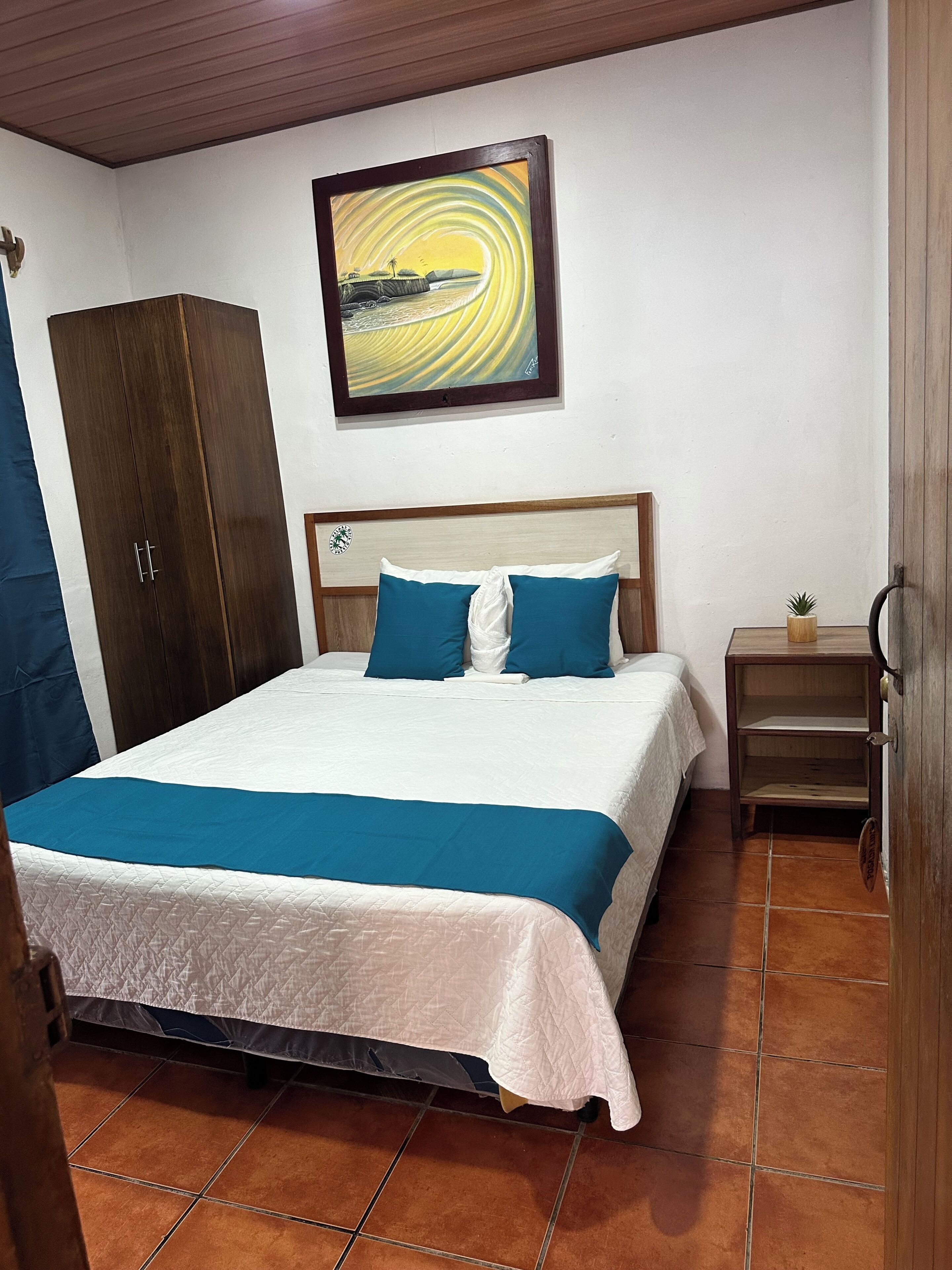 A bedroom with a bed, a wooden wardrobe, a nightstand, and a wall-mounted painting.