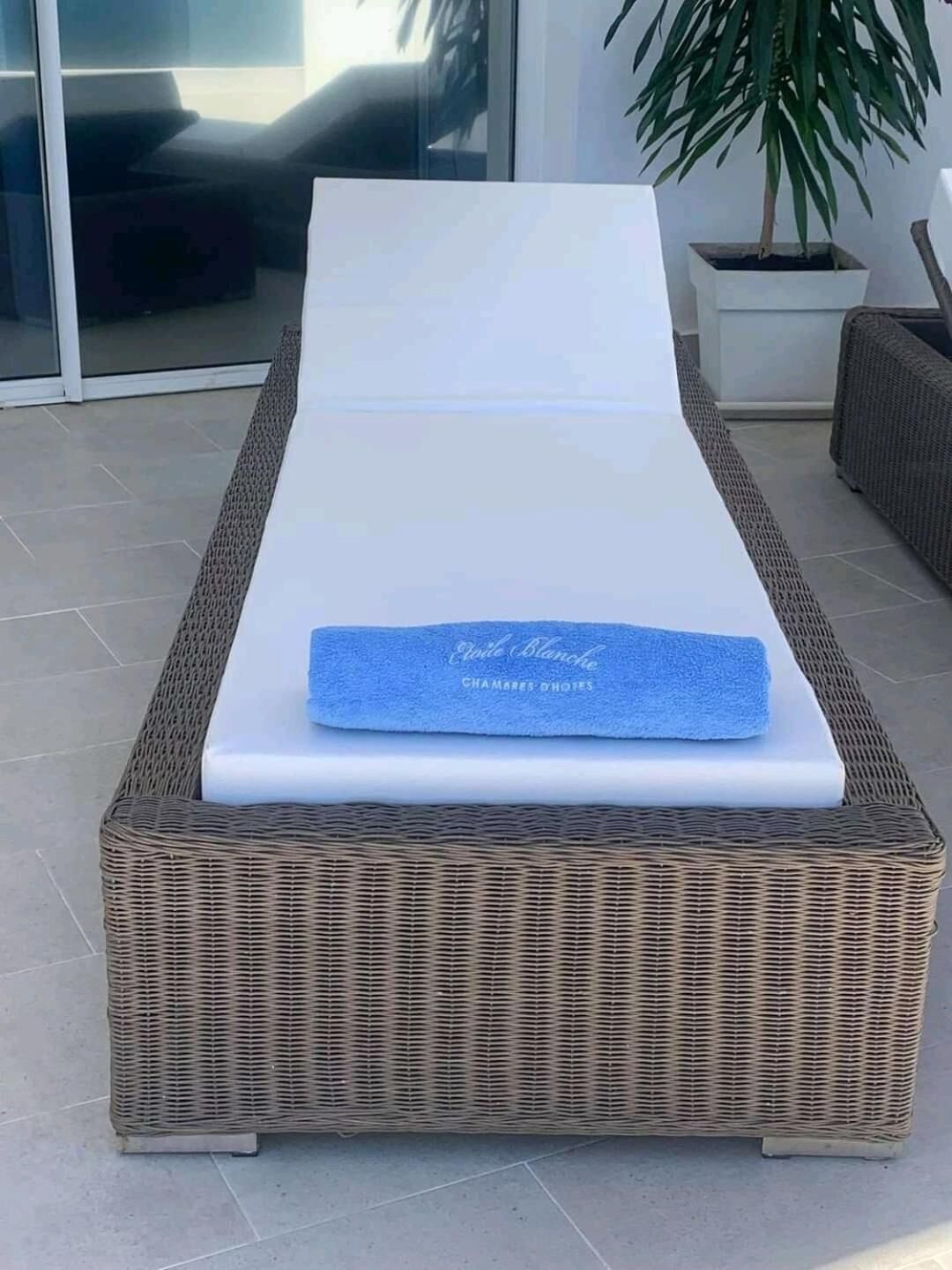 Un fauteuil de relaxation en osier, doté d’un matelas blanc et d’une serviette bleue, est placé sur une terrasse carrelée.