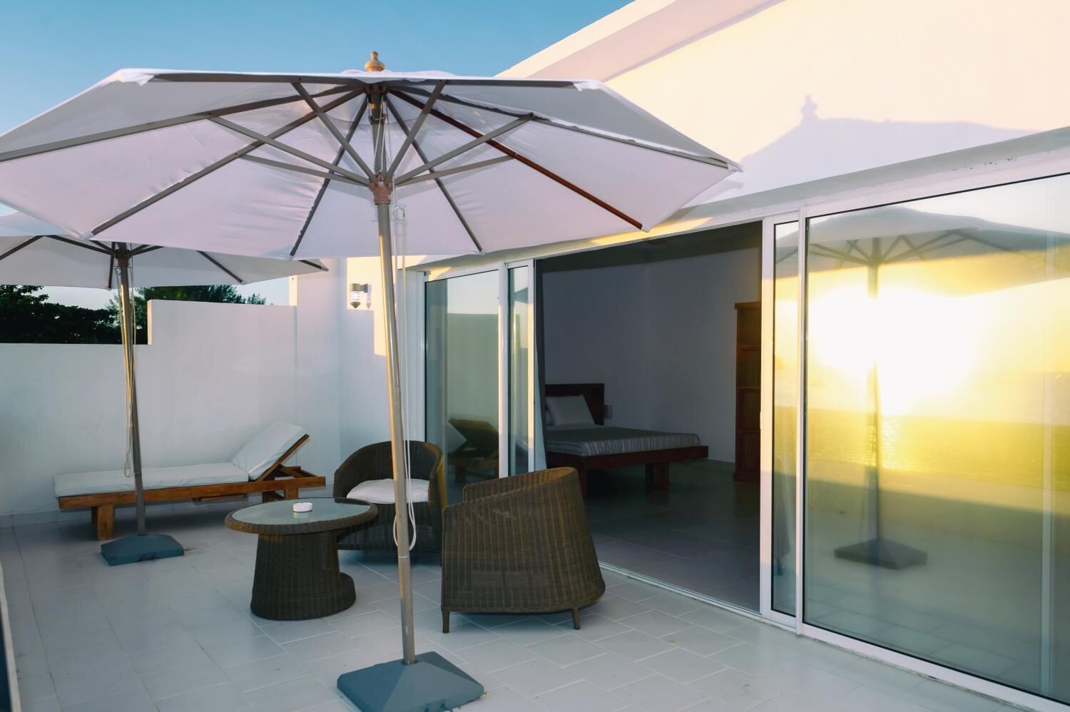 A modern outdoor patio with a glass door leading to a bedroom, a white umbrella, wicker furniture, and a small table.