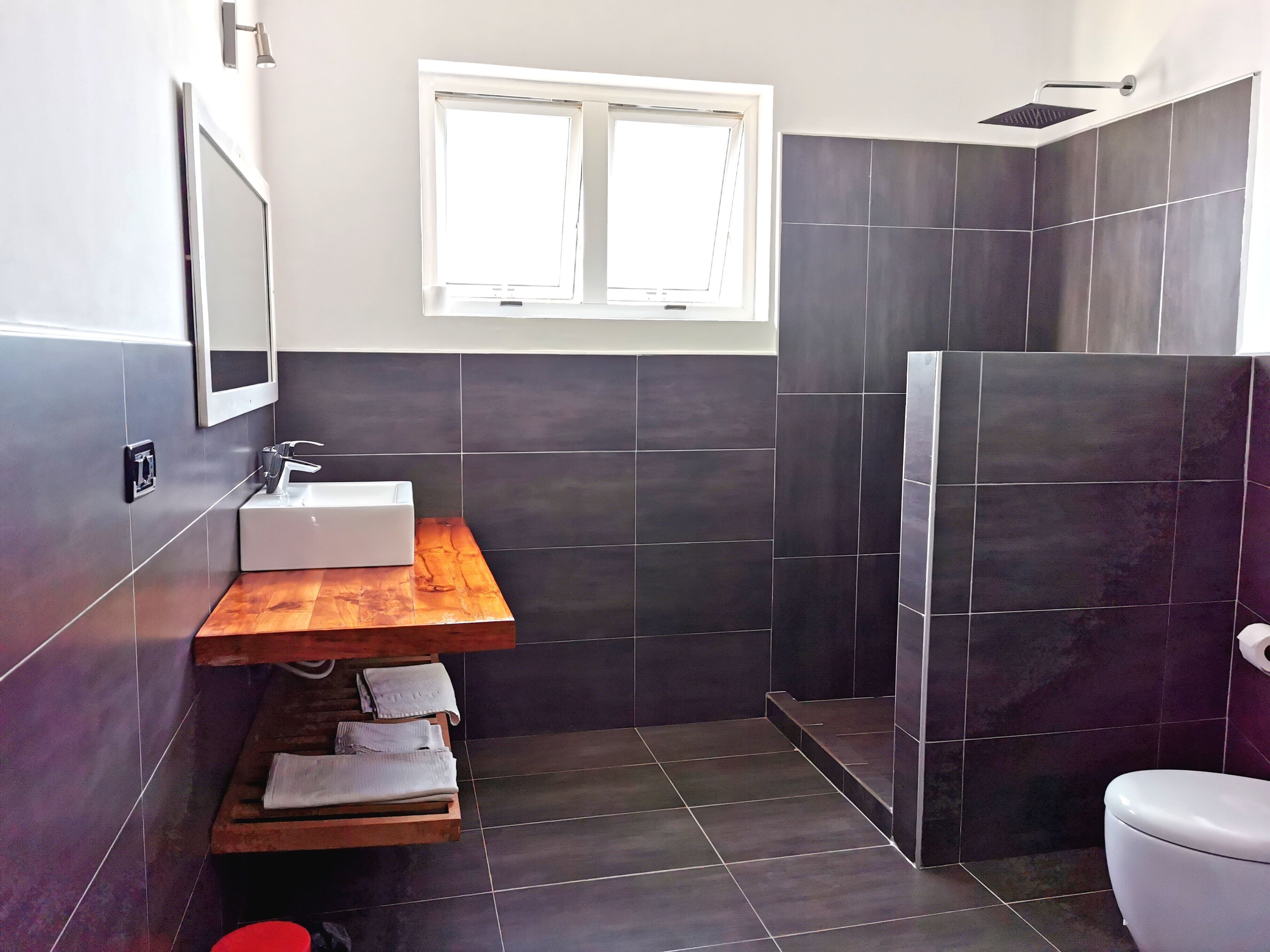 A modern bathroom with a wooden vanity, a white sink, a mirror, a window, and a shower area.