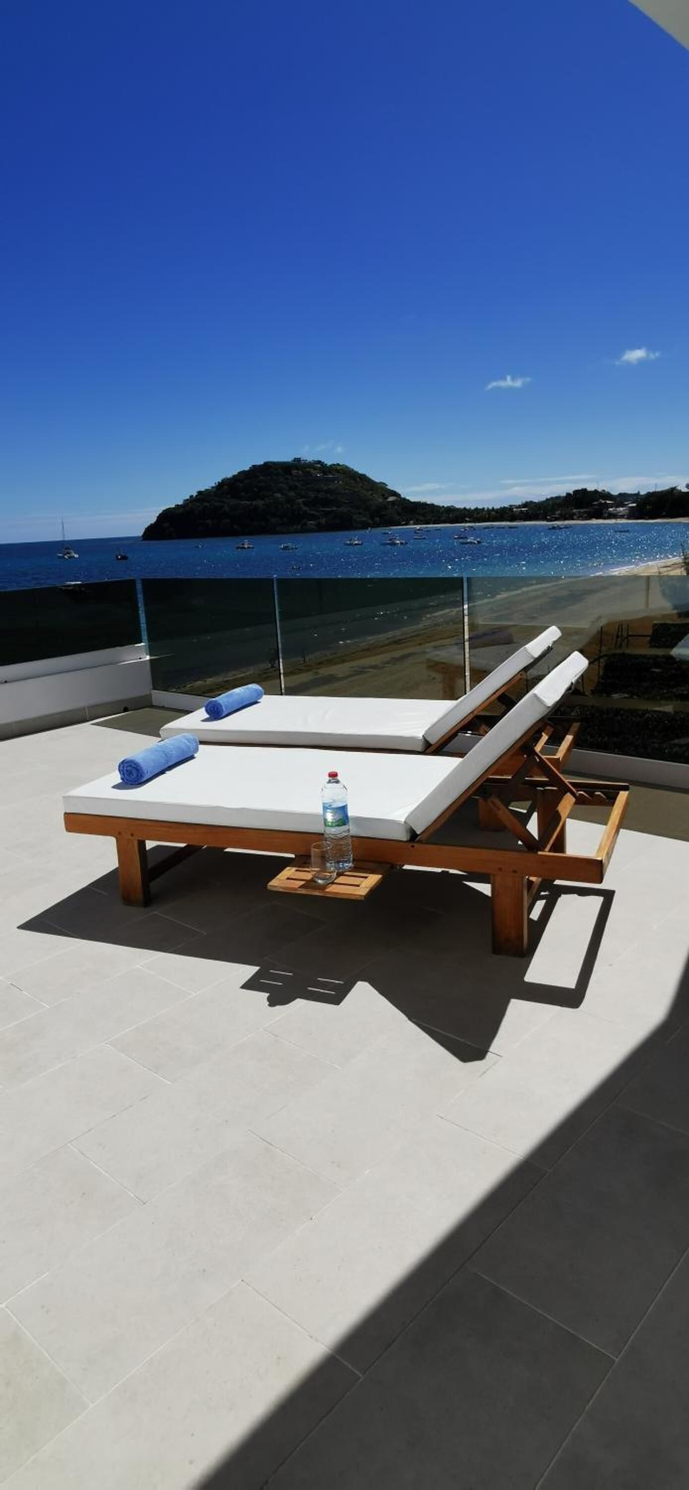 A sun lounger with a white cushion and a blue towel, a bottle of water, and a glass on a tiled patio overlooking a beach with boats and a small island.