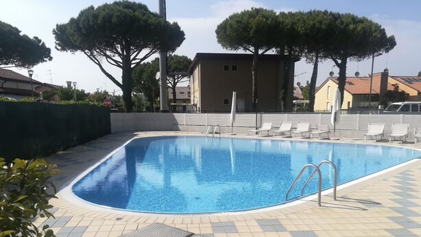Ein rechteckiger Swimmingpool mit Fliesen umgeben, mit Liegestühlen und Bäumen gesäumt.