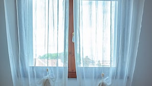 Ein Hotelzimmer mit Bett, weißen Vorhängen und einem Fenster mit Blick auf Gebäude.