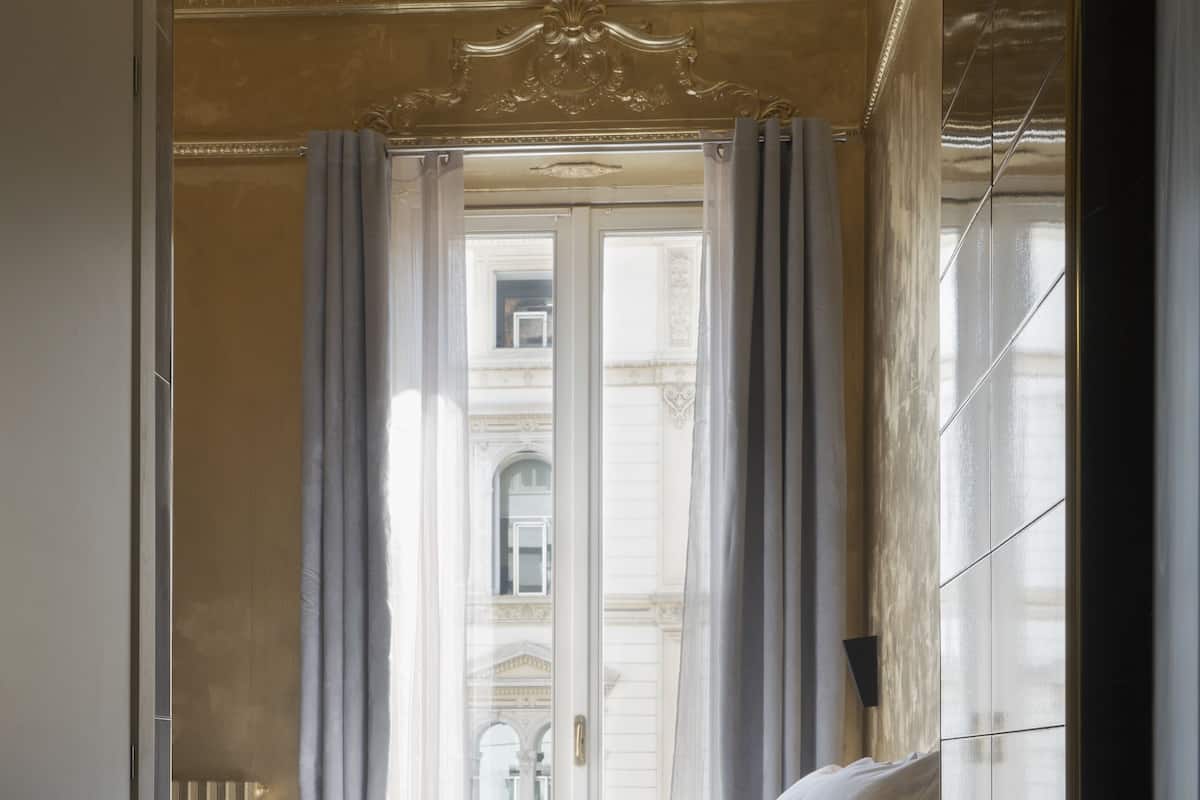 A hotel room with a checkered floor, a bed with pillows, a window with curtains, and a view of a building outside.