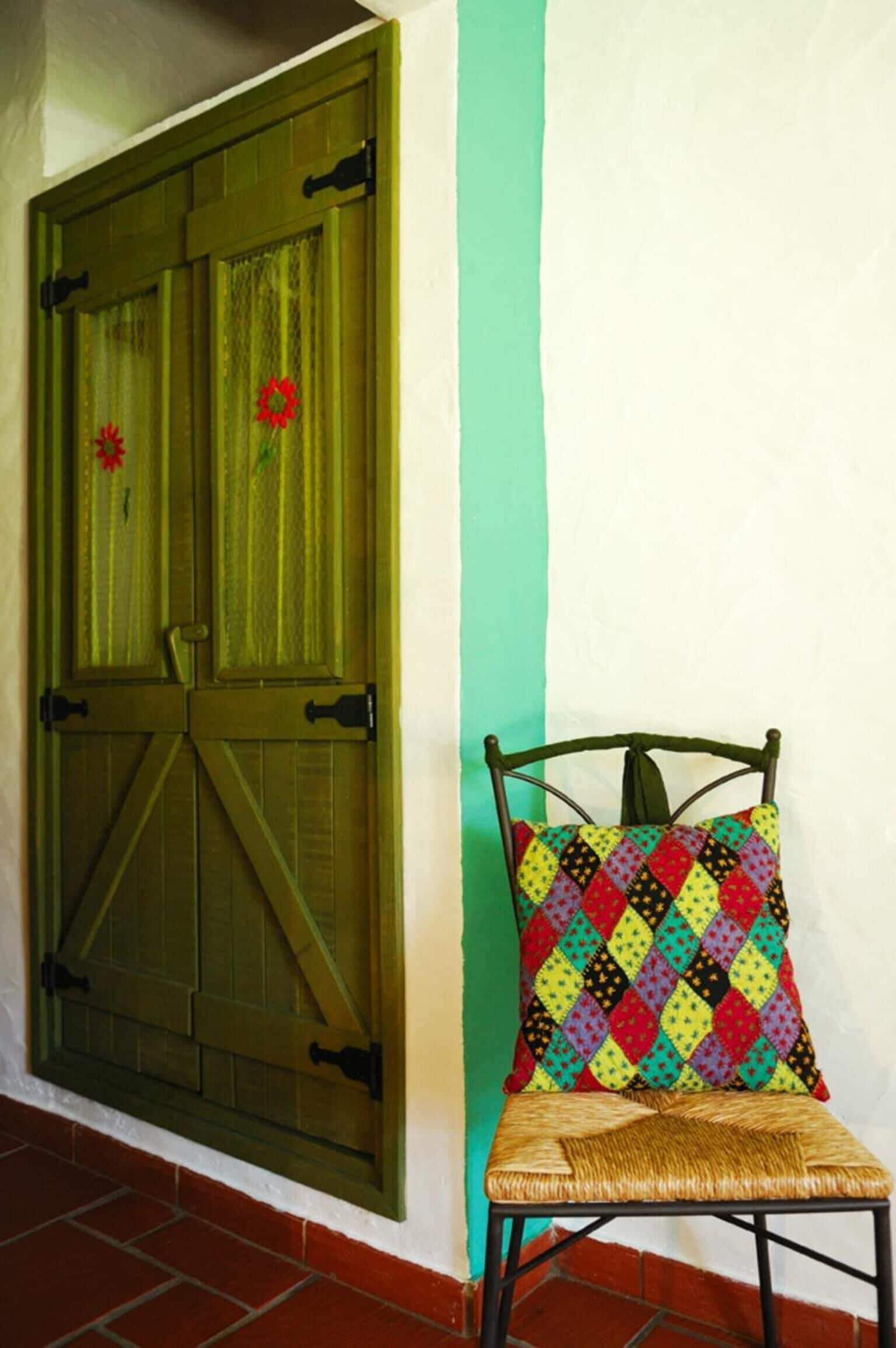 A chair with a colorful quilted pillow against a wall with a green door.
