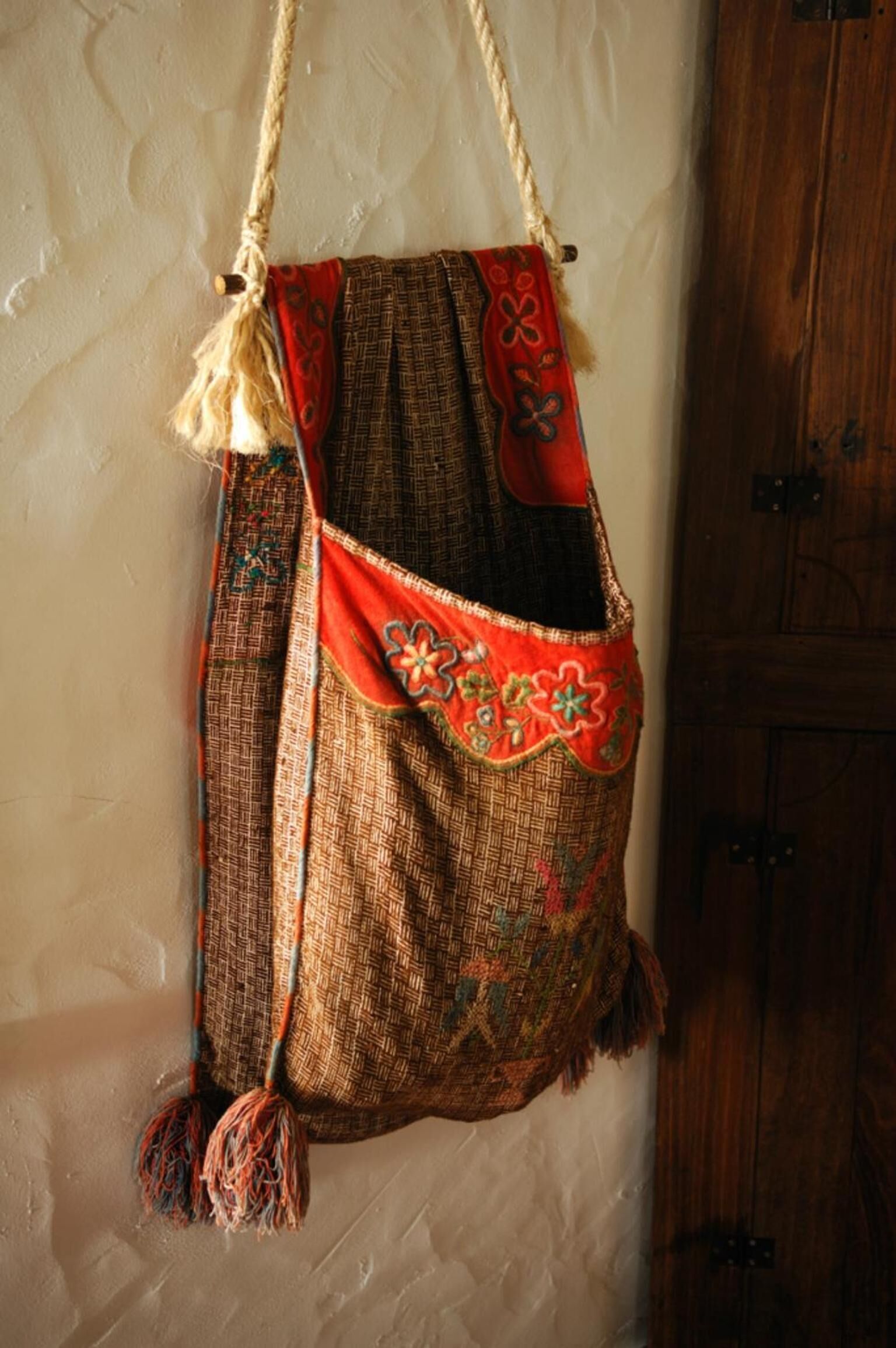 Un bolso tejido con flecos y diseños decorativos, colgado de una cuerda frente a una pared.