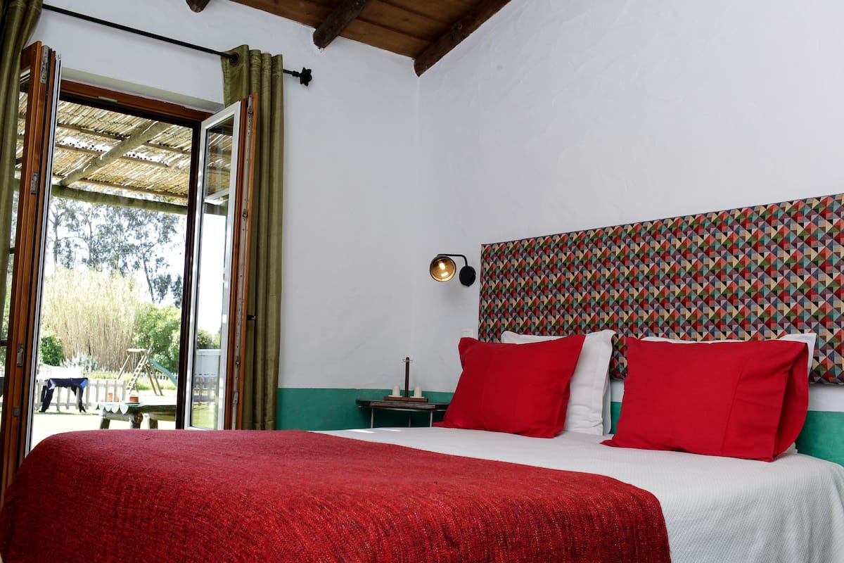 A bedroom with a large bed, red and white bedding, a patterned headboard, a nightstand with a lamp, and a view of the outdoors through an open door.
