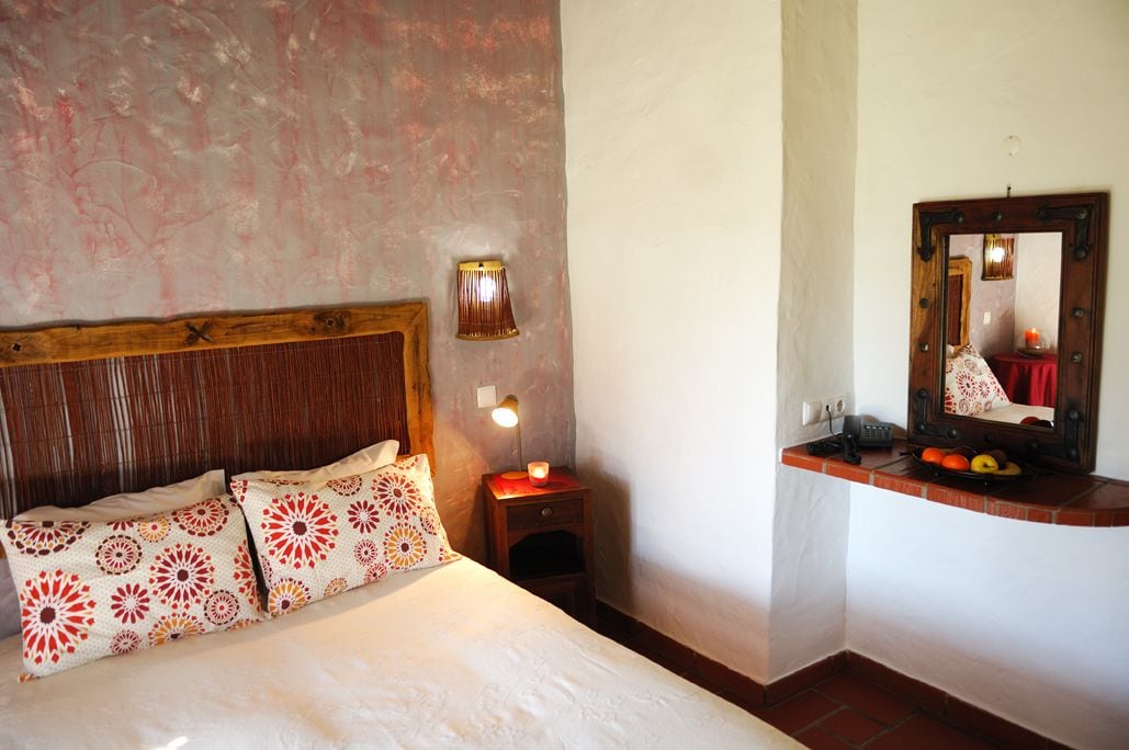A bedroom with a wooden headboard, a bedside lamp, a mirror, and a small table with fruits.